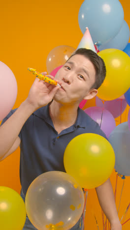 Vertical-Video-Studio-Portrait-Of-Man-Wearing-Party-Hat-Celebrating-Birthday-With-Balloons-And-Party-Blower